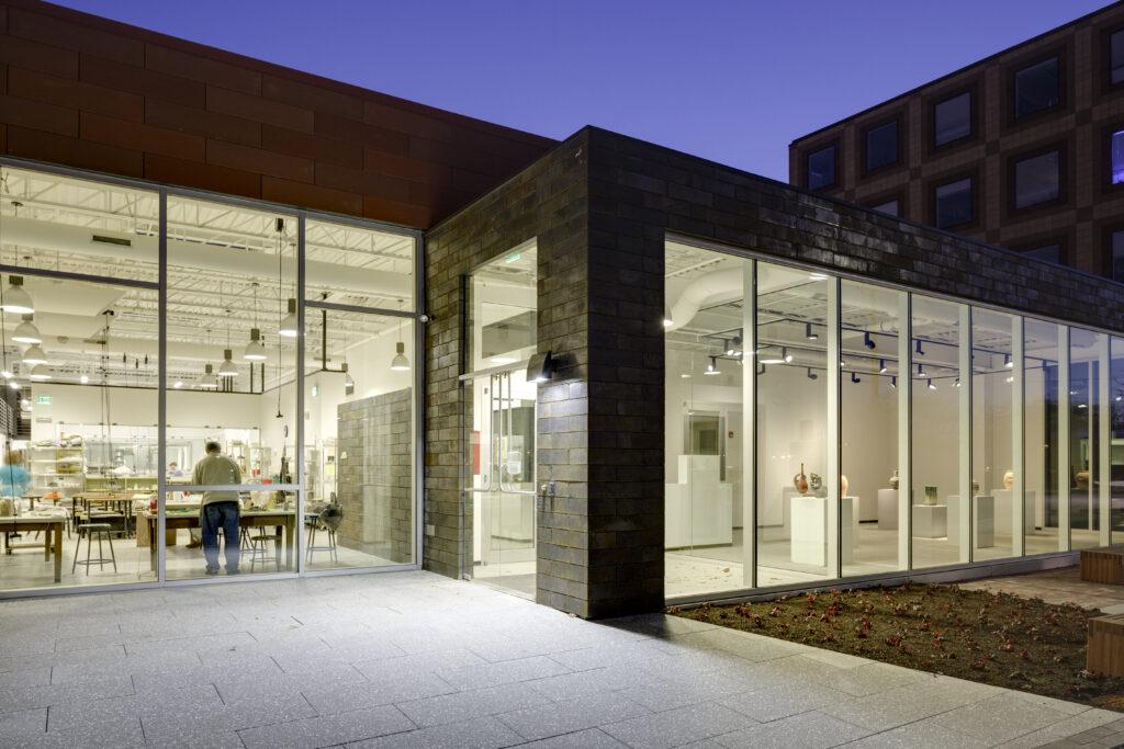 Modern glass-walled building at dusk with people working at tables inside under hanging lights.