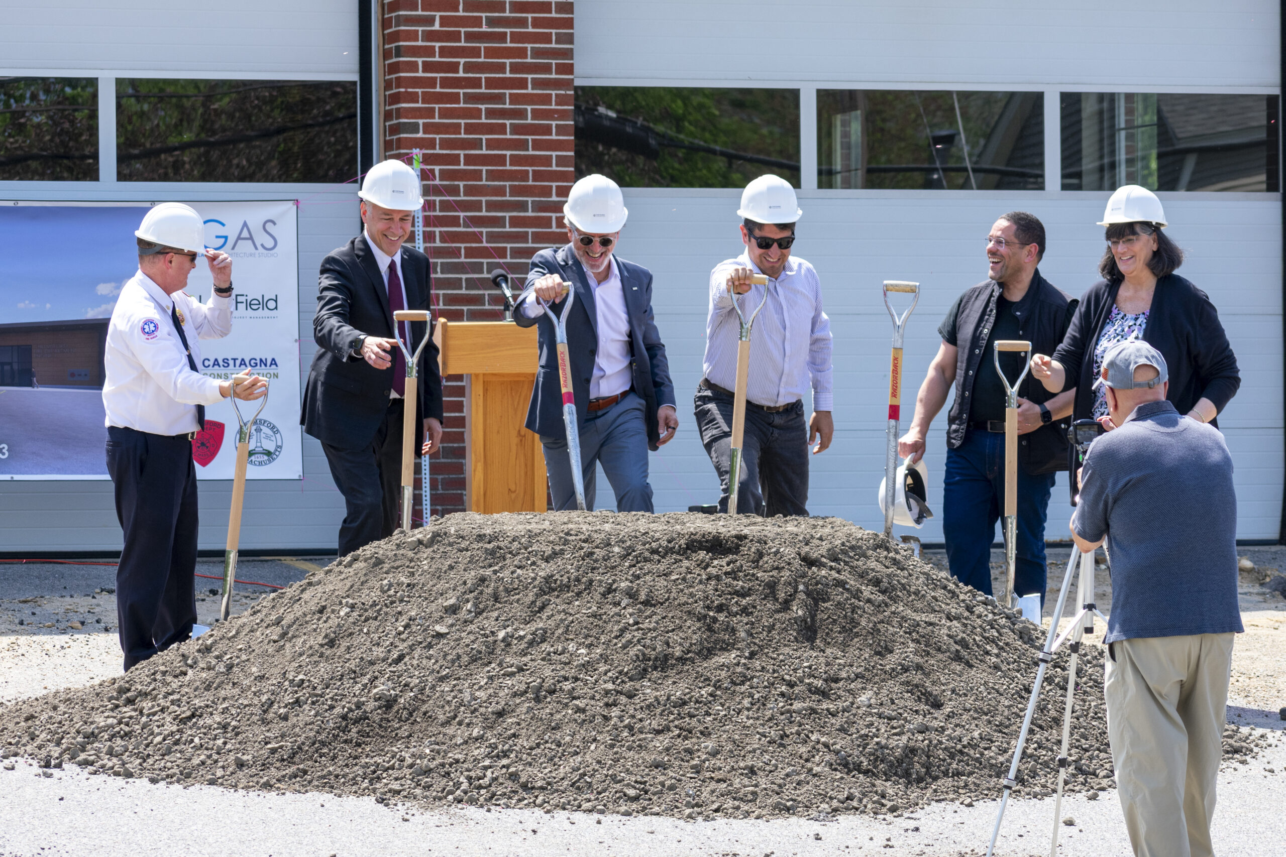 Group of professionals in hard hats at a building's groundbreaking ceremony, shovels in hand around a dirt pile.