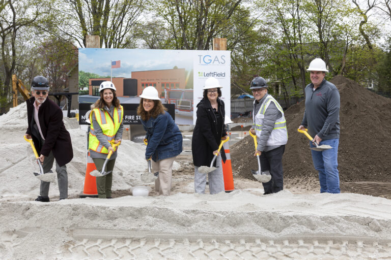 Group of construction workers and engineers with shovels at a site, a large sign behind them shows TGAS and LeftField in the background.
